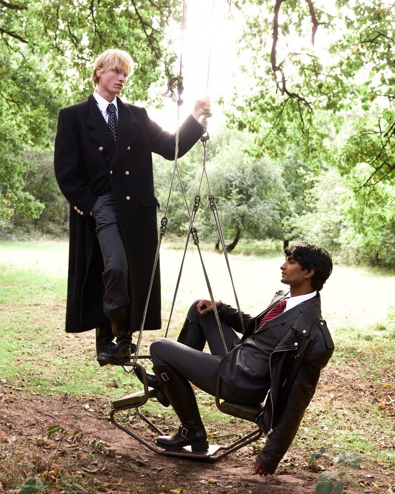 Two male fashion models in a forest setting. One model is standing, wearing a black coat, while the other is sitting on a swing.