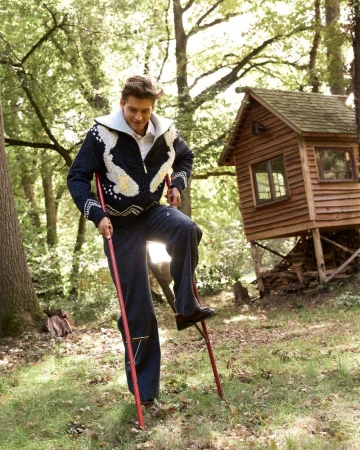 Man balances on stilts in a sunlit forest, wearing a patterned sweater and dark trousers, a wooden cabin on stilts is visible in the background.