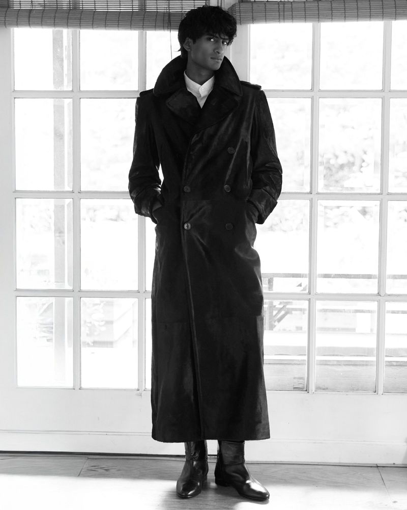 Full-length portrait of a young adult man wearing a long black coat and dress shoes, posed in front of a window with white trim.