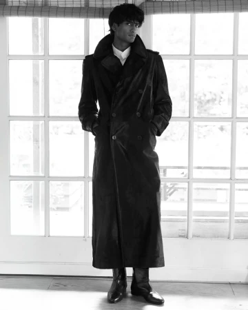 Full-length portrait of a young adult man wearing a long black coat and dress shoes, posed in front of a window with white trim.