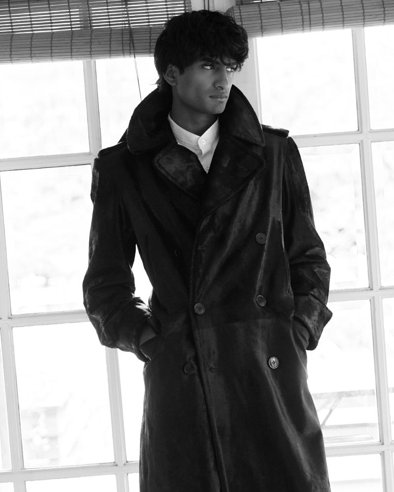 Black and white portrait of a young adult man wearing a long, dark coat, standing near a window, looking to the side in a studio setting.