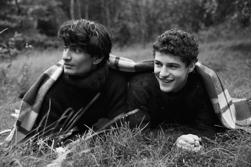 Two young adult men smile while laying in grass, sharing a checkered blanket. They are wearing sweaters in a black and white outdoor shot.