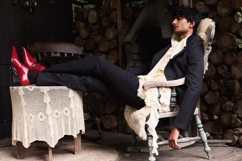 Man in a black suit lounging in a wooden chair on a porch. He is wearing bright red shoes. Logs are stacked against the house wall.