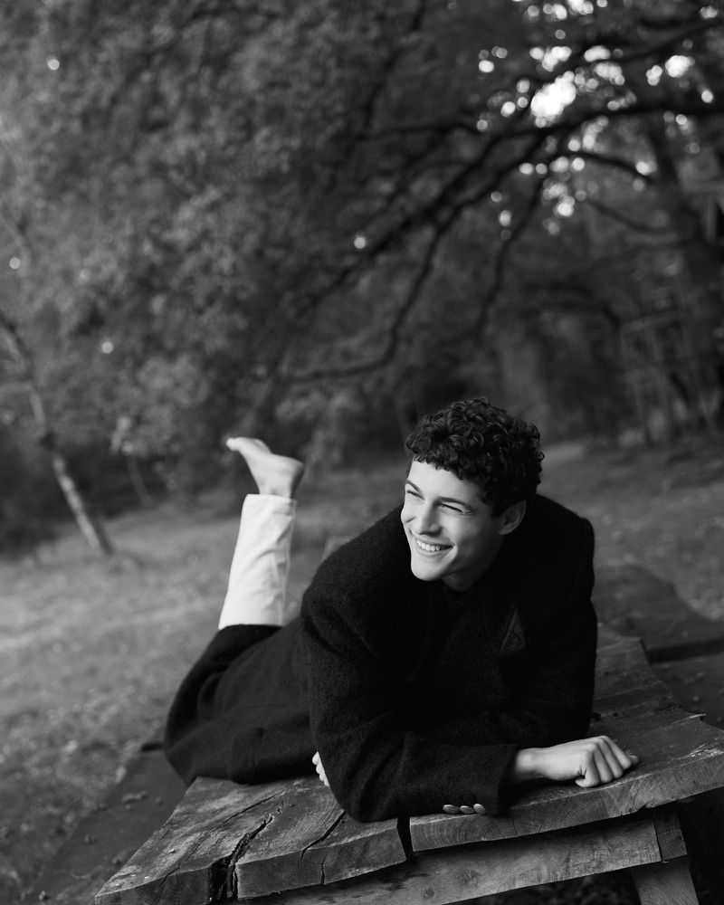 Monochrome portrait of a smiling young man lying on a wooden table outside in a coat, with trees and greenery in the background.