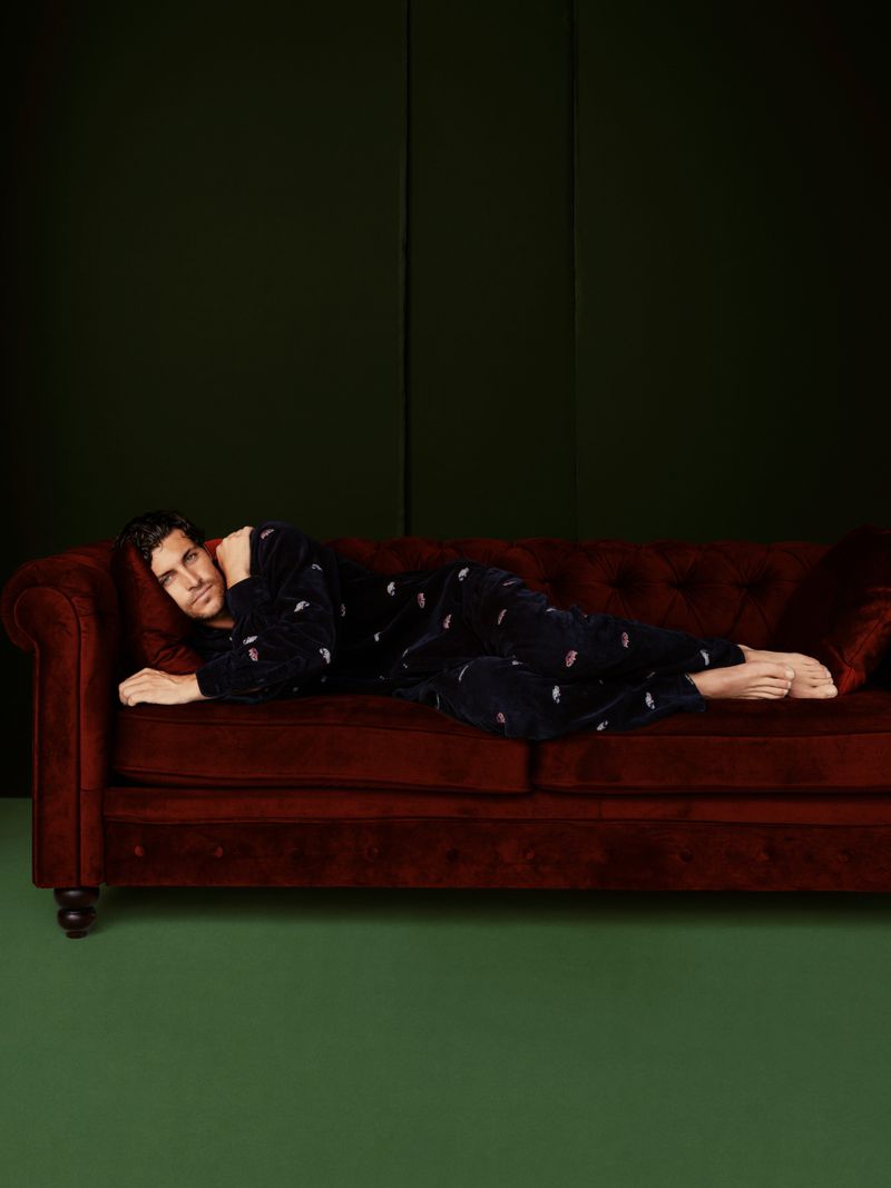 Adult man lying on a red velvet couch wearing dark pajamas in an indoor setting with a green wall in the background.
