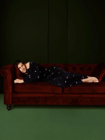 Adult man lying on a red velvet couch wearing dark pajamas in an indoor setting with a green wall in the background.