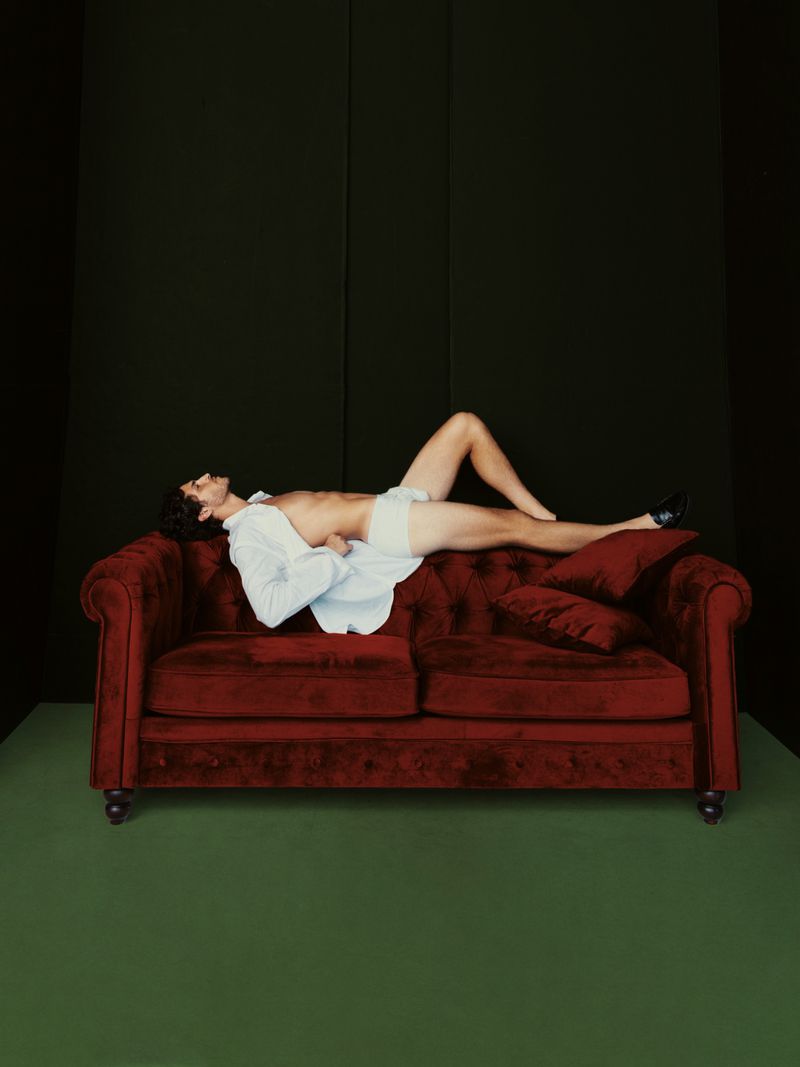 Man in a white shirt and briefs lies on a red velvet couch in a dark room with a green floor, showcasing relaxation and modern style.