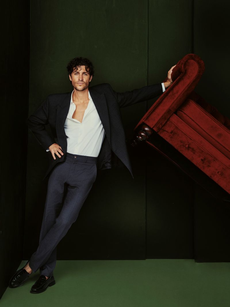 Man in black blazer and blue pants leans against a red chair in a photo studio with a green background, striking a pose.