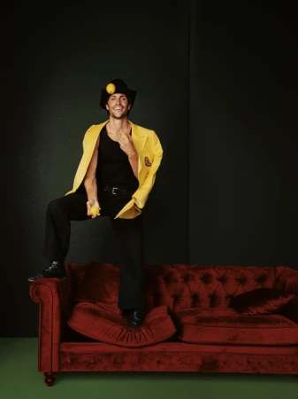 A stylish man in a black hat and yellow jacket playfully poses on a red tufted couch, balancing lemons in his hand and on his head.