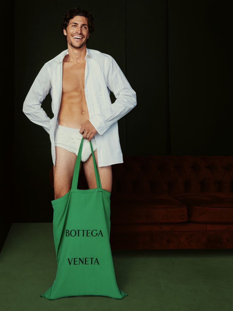 Smiling man poses indoors with Bottega Veneta tote bag and unbuttoned shirt, showing torso. Dark background with sofa.
