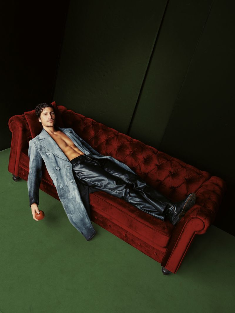 Adult male lounging on a red velvet couch in a photo studio, wearing a blue denim coat and black leather pants with an apple nearby.