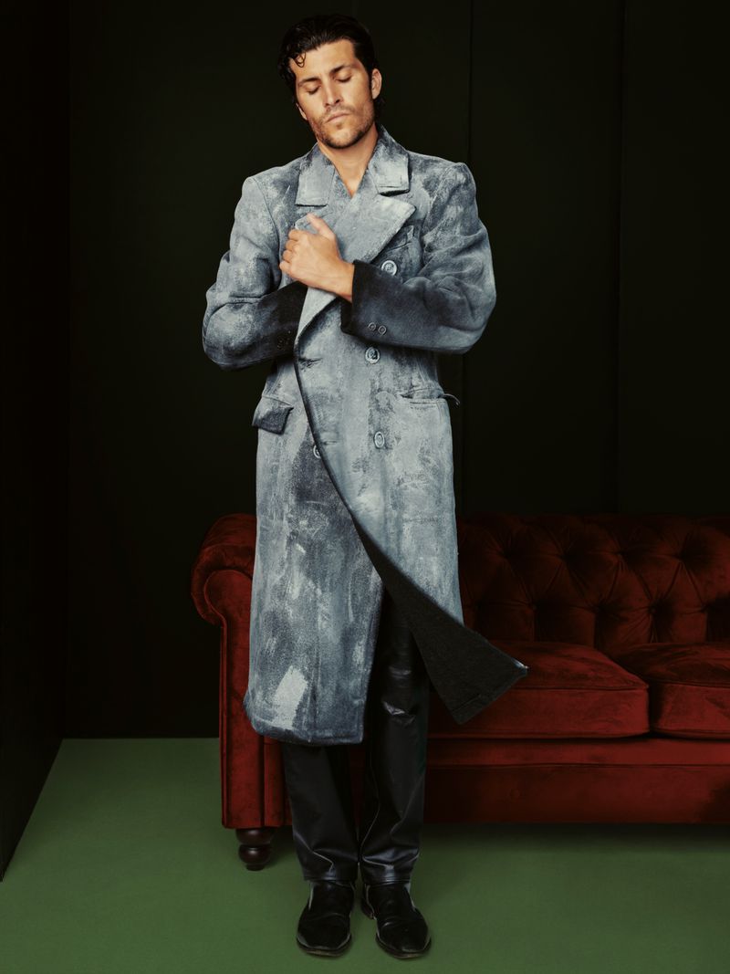 Adult male model poses indoors with a painted blue trench coat, dark trousers, and red velvet sofa in a dark room with green floor.