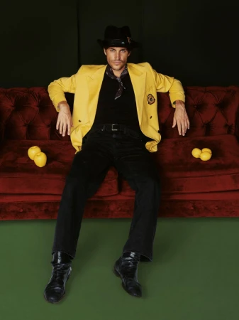 A man in a yellow jacket and black hat sits on a red velvet couch with lemons in a studio setting with green flooring and a dark background.