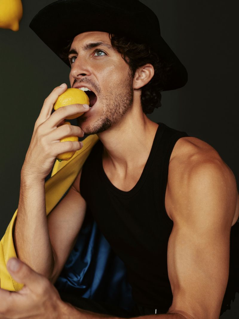 Man wearing a black hat and tank top is biting into a lemon, holding another lemon in his hand. Yellow and blue fabric on shoulder.