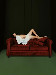 Man in an open white shirt and briefs reclining across the top of a deep red velvet chesterfield sofa against a dark background.