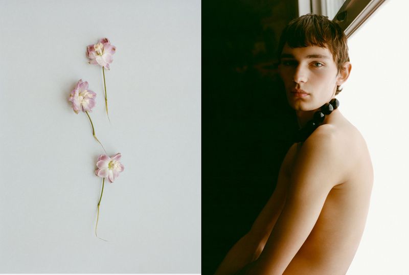 Portrait of a shirtless male model with a beaded necklace alongside three pink flowers on a two-tone light and dark background.