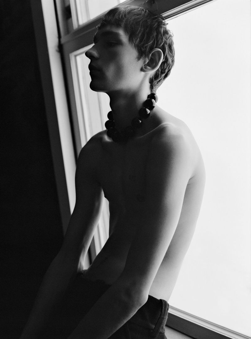Monochrome portrait of a young adult male posing shirtless by a window with a dark beaded necklace, captured in a dramatic, close-up shot.