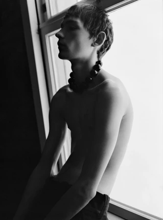 Monochrome portrait of a young adult male posing shirtless by a window with a dark beaded necklace, captured in a dramatic, close-up shot.