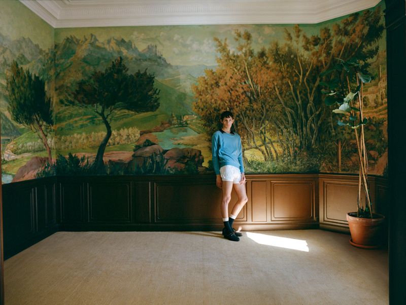 Woman in blue top and white shorts poses in a room with a mural of a mountain landscape. A potted plant stands nearby.