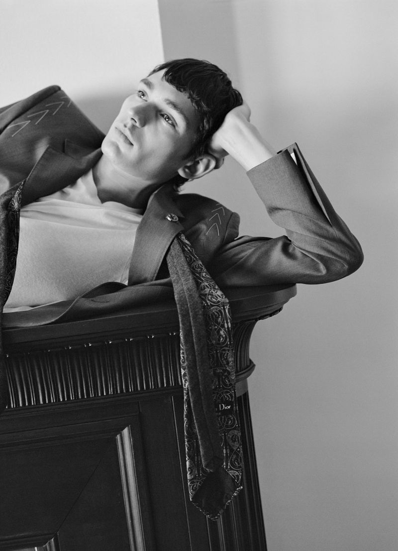 Black-and-white portrait of a male model in a suit reclining against a fireplace, looking to the side with a serious expression.