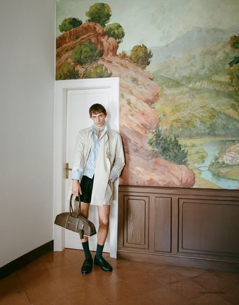 A slender man wearing a trench coat, blue shirt, and black shorts poses indoors while holding a satchel bag in a full shot.