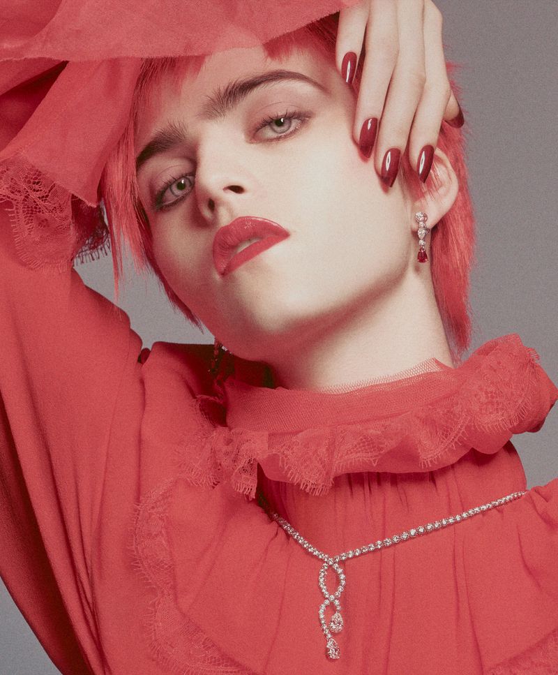 Adult woman with short red hair and red lipstick poses wearing matching red dress and diamond jewelry in a studio setting.