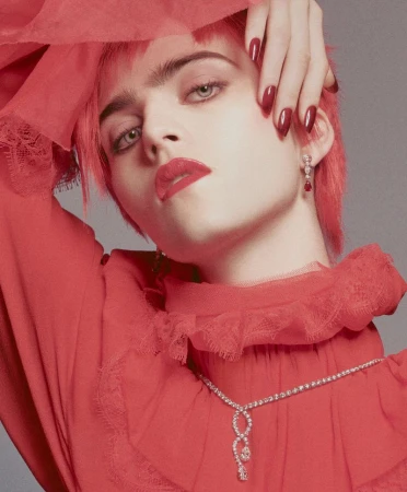 Adult woman with short red hair and red lipstick poses wearing matching red dress and diamond jewelry in a studio setting.