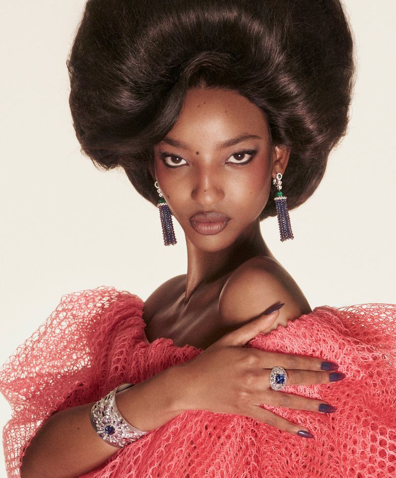 Portrait of an adult female with large afro hairstyle, wearing a pink lace dress, large jewelry, and dark nail polish against white.