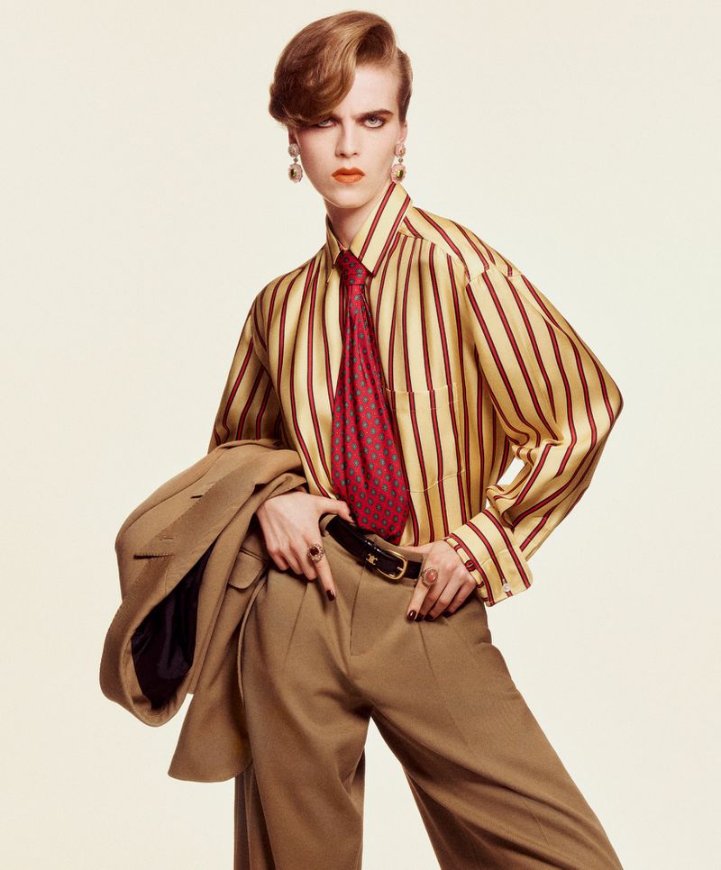 Fashionable adult poses indoors wearing a striped shirt, tie, and pants, holding a tan suit jacket, showcasing a modern retro aesthetic.