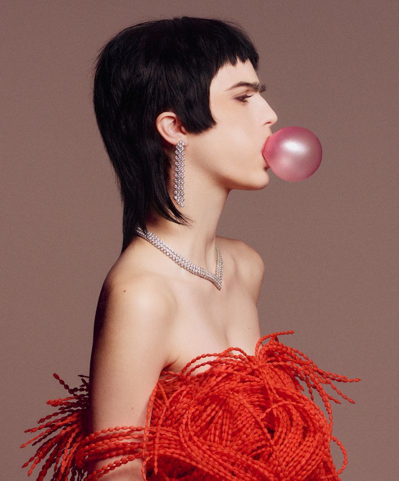 Side-profile portrait of an adult female with a short black mullet blowing a pink bubble, wearing jewelry and a red feathered top.