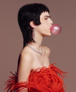 Model with dark mullet blowing a pink bubblegum bubble wearing diamond earrings and a textured red top