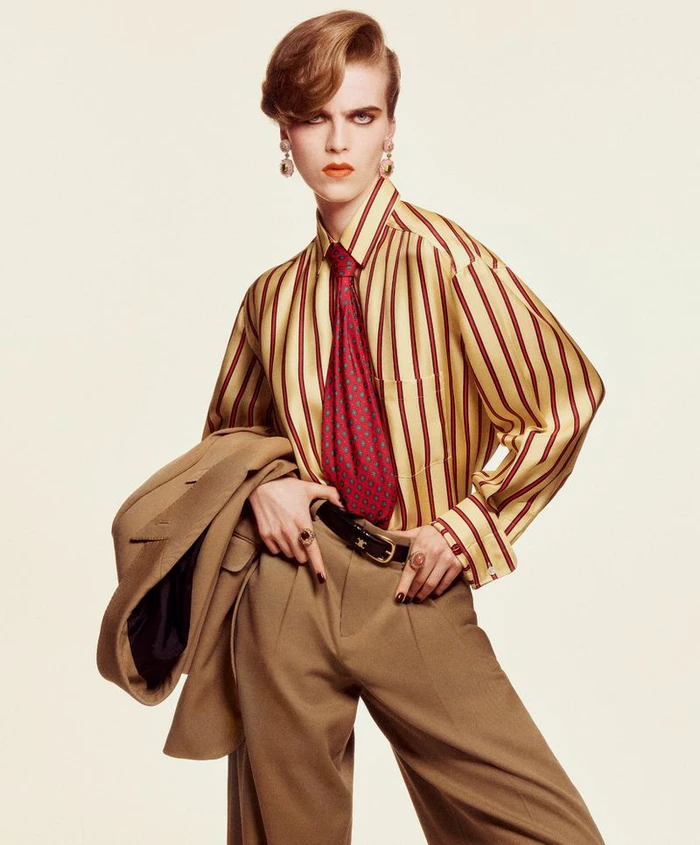 Model Libby Bennett posing in a gold and red striped shirt, red patterned tie, and camel trousers against a light seamless backdrop.
