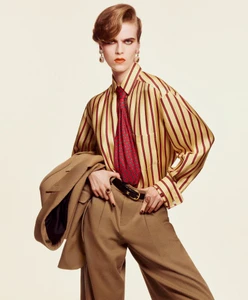 Model Libby Bennett posing in a gold and red striped shirt, red patterned tie, and camel trousers against a light seamless backdrop.
