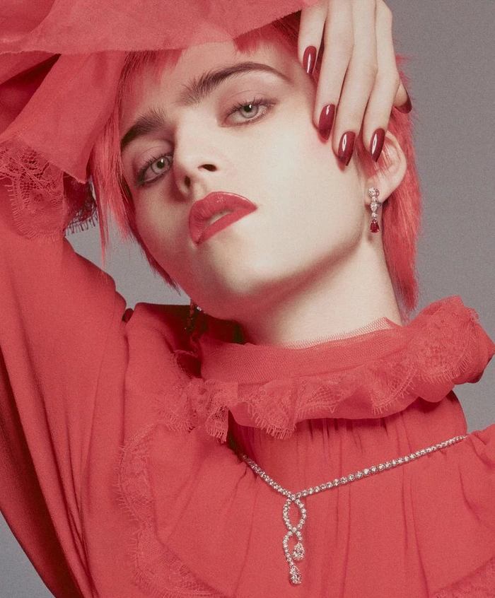 Close-up portrait of a model with red hair wearing a red high-neck lace blouse, diamond necklace, and red manicured nails against a grey backdrop.