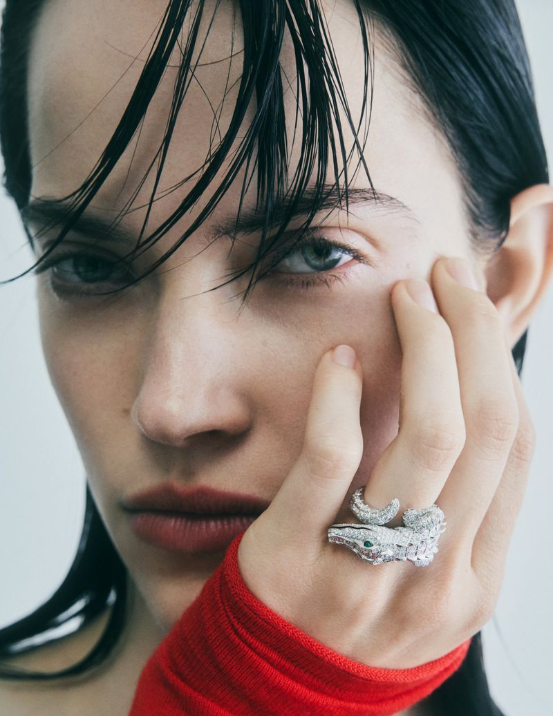 Close-up studio shot of a brunette woman with blue eyes, wearing a snake ring and a red top against a grey studio backdrop.
