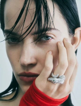 Close-up studio shot of a brunette woman with blue eyes, wearing a snake ring and a red top against a grey studio backdrop.