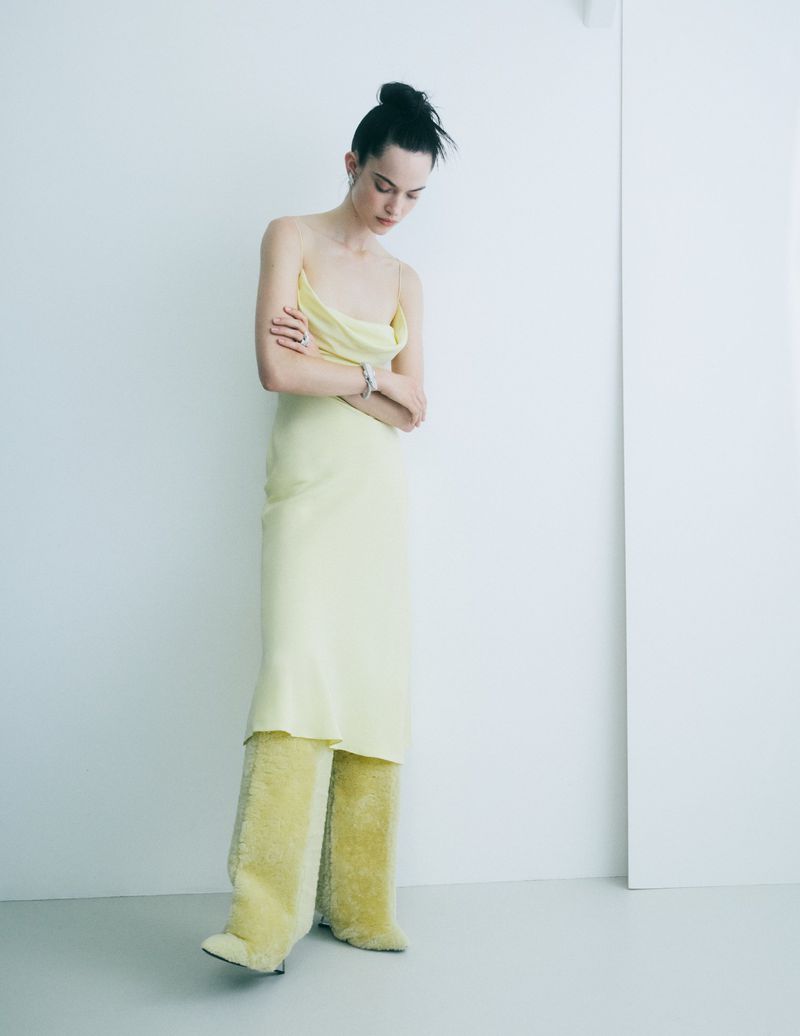 A woman in a pale yellow dress and furry pants stands indoors against a white wall, looking down with her arms folded.