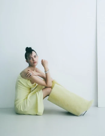 Model seated on studio floor wearing a pale yellow slip dress and oversized textured boots with diamond jewelry.