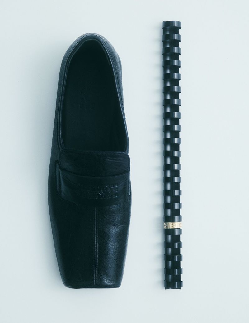 Top-down shot of a black leather loafer paired with a geometric black tube against a stark white background for fashion editorial.