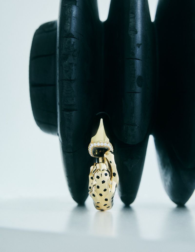 High-angle studio shot of a gold, hand-shaped earring with black spots hanging against a textured black background. Fashion still life.