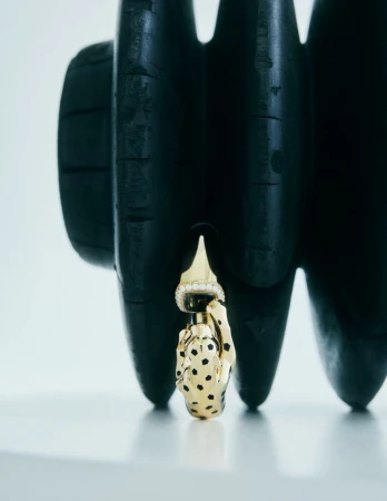 High-angle studio shot of a gold, hand-shaped earring with black spots hanging against a textured black background. Fashion still life.