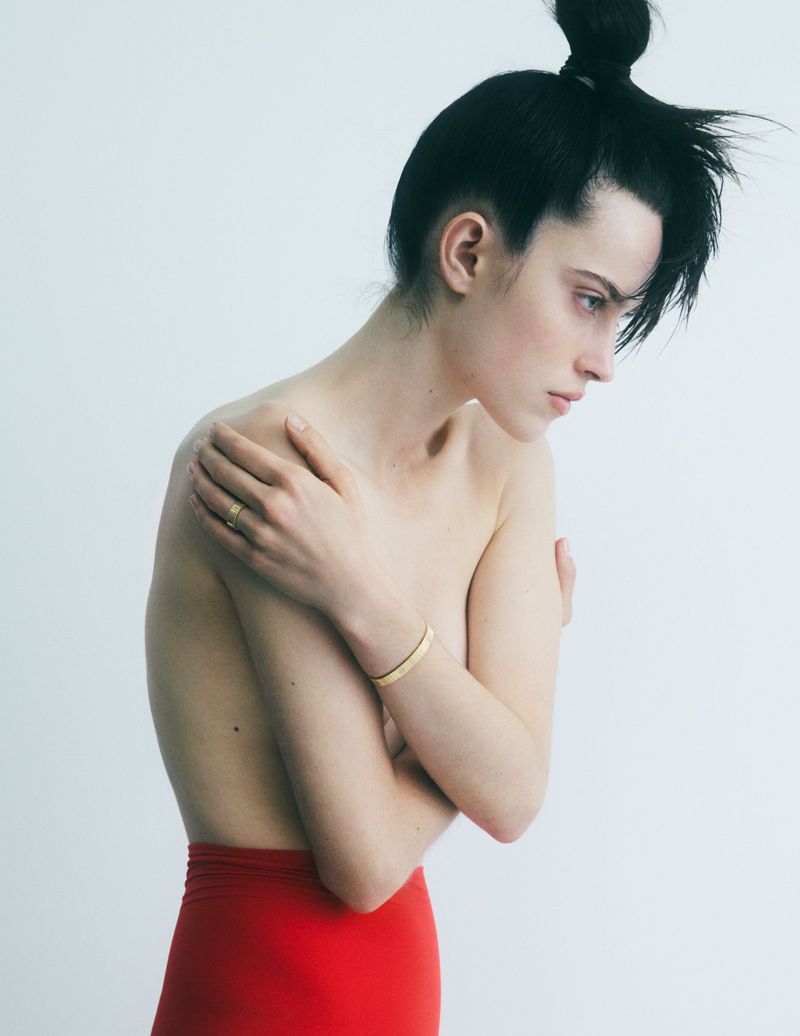 Adult woman in red skirt and stylish hair bun posing topless in a minimalist white studio background with gold ring and bangle.