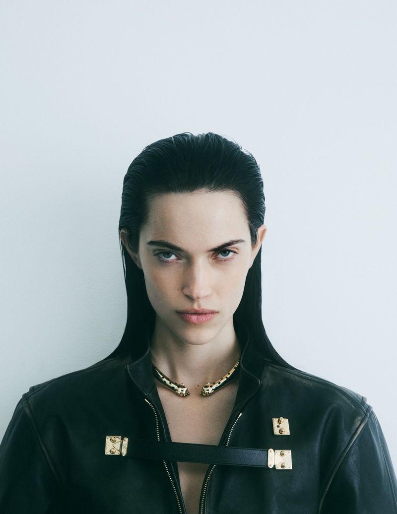 Close-up studio shot of a young adult woman with straight, dark hair, wearing a black leather jacket and a gold necklace.