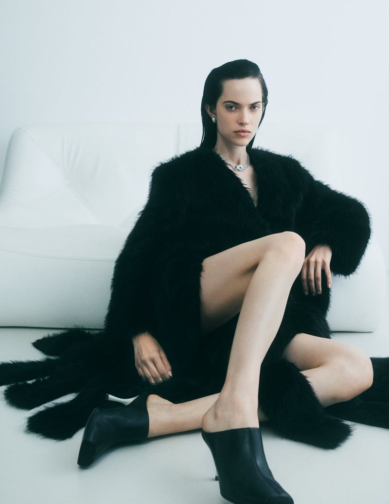 Brunette woman in fur coat and black mules sits on floor against white couch, wearing a necklace and earrings in a fashion studio setting.