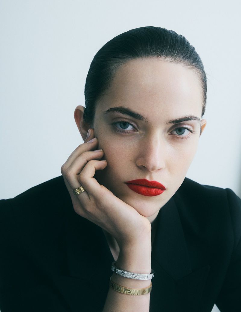 Close-up portrait of a woman resting her chin on her hand, wearing fine jewellery bracelets, a ring, and red lipstick against a white background.