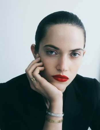 Close-up portrait of a woman resting her chin on her hand, wearing fine jewellery bracelets, a ring, and red lipstick against a white background.