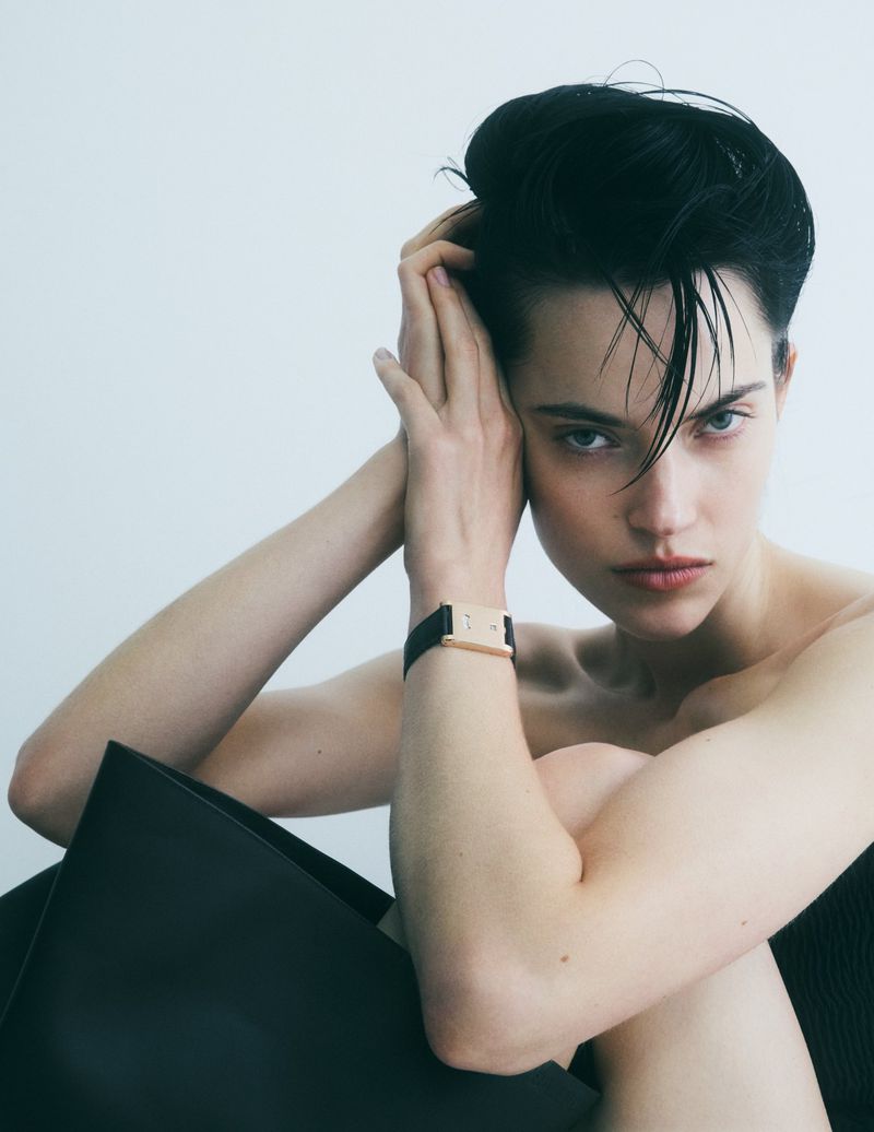 Portrait of a young adult woman with slicked-back dark hair in a minimalist studio setting, wearing a black wristwatch and posing intimately.