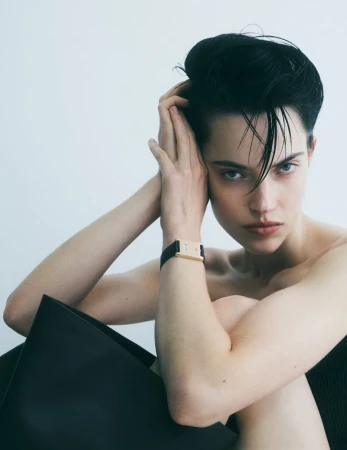 Portrait of a young adult woman with slicked-back dark hair in a minimalist studio setting, wearing a black wristwatch and posing intimately.