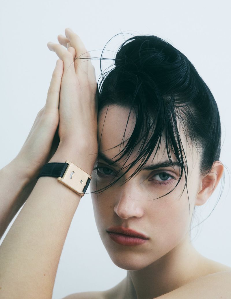 Close-up fashion shot of a brunette woman with her hands up, wearing a gold and black wristwatch, posing in front of a white background.