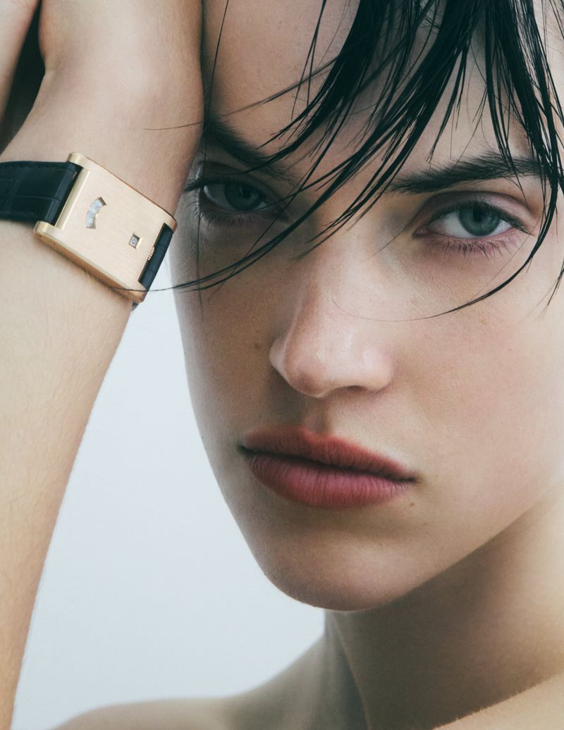Close-up of a brunette woman's face, she wears a black and gold wristwatch, posing against a light backdrop, showing a beauty and fashion shot.